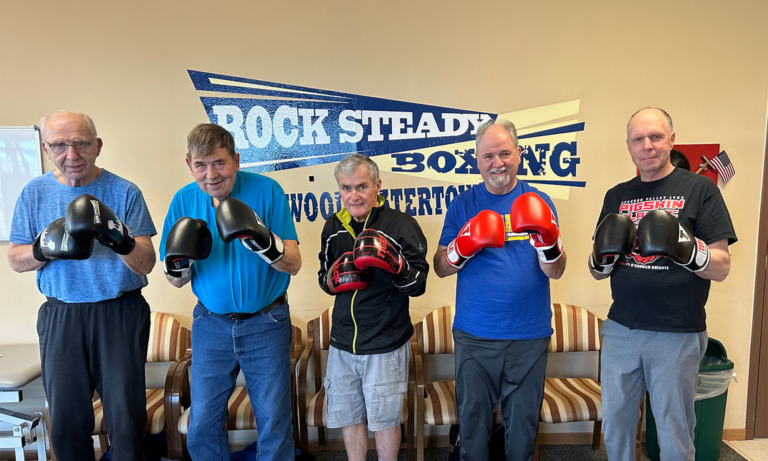 Five senior men stand in front of a “Rock Steady Boxing” sign, posing with boxing gloves. The group smiles proudly in a boxing gym, suggesting participation in a fitness or boxing program.